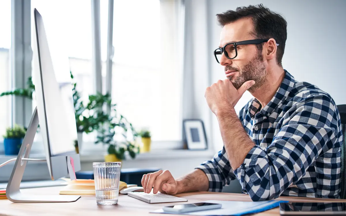 A man with glasses sits at his desk in front of the screen and thinks
