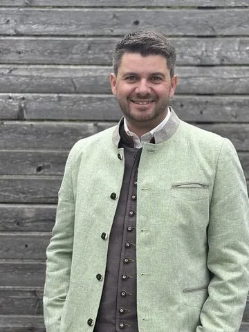 Philipp vor einer Holzwand, lächelnd, im grünen Janker